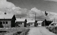 Old black-and-white photo, five wooden houses along a small gravel street, Finnish flag up in a flagpole.
