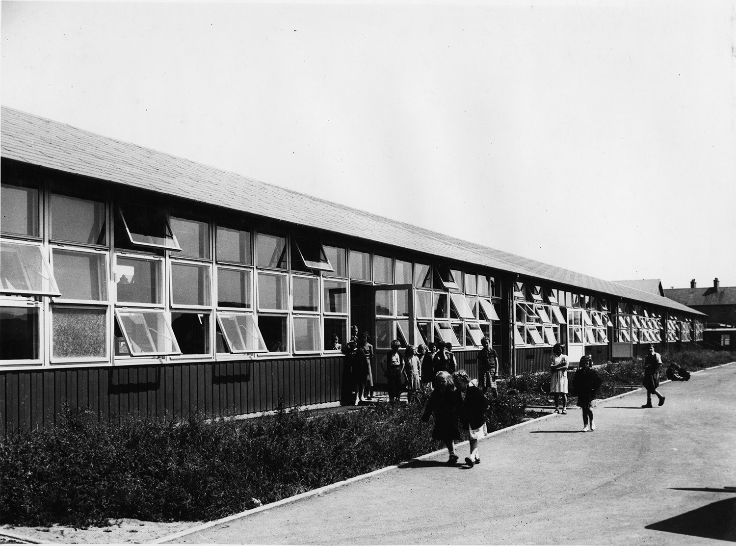 An old black and white photo of a Puutalo wooden school. The school's wall has many windows right next to each other and some of them are open.