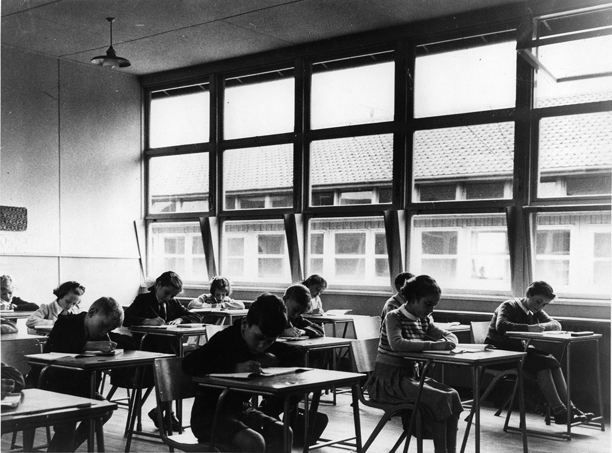 An old black and white picture of a classroom, children writing at their desks, a wall of windows in the background.