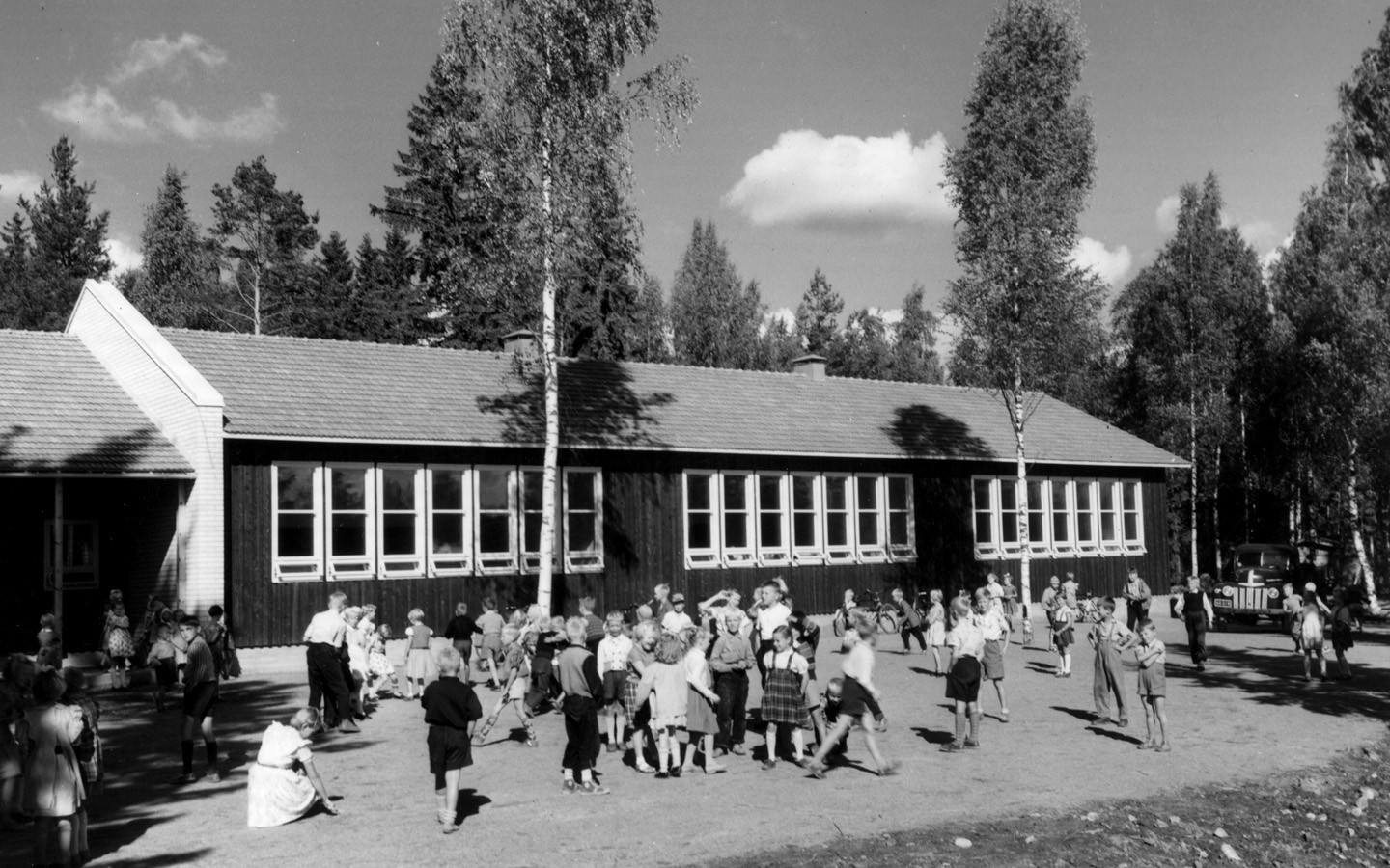 An old black and white picture of a wooden school where there are children playing in the yard.