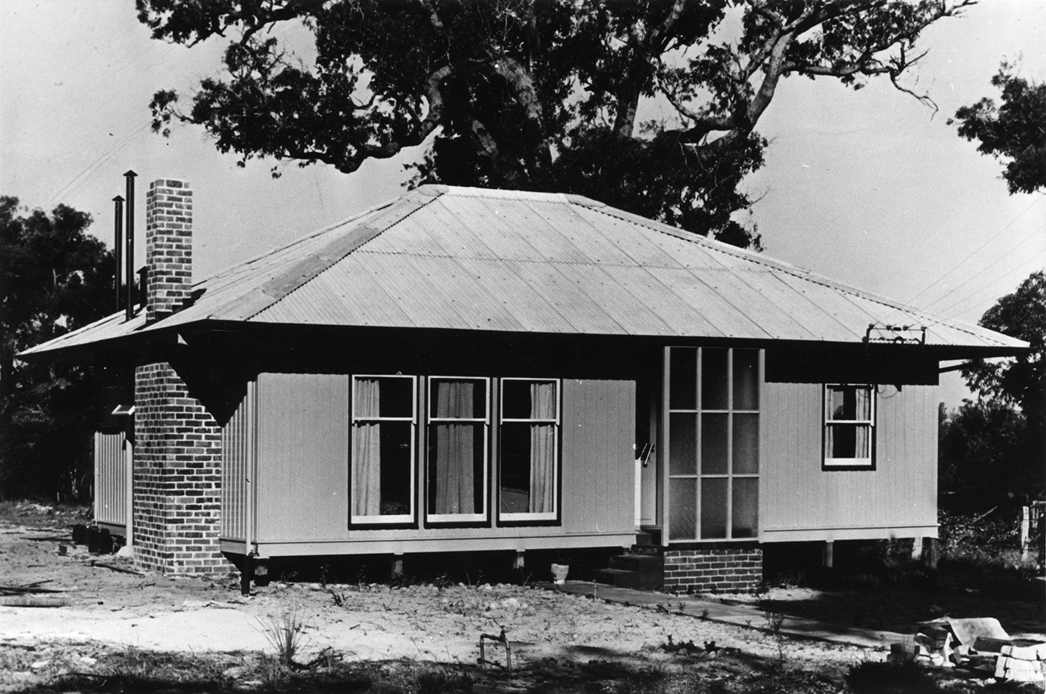 A black and white photo of a Puutalo Oy’s wooden house. The eaves of the house are long, the windows are relatively large and on the side wall there is a chimney. In the background is a large and winding tree.