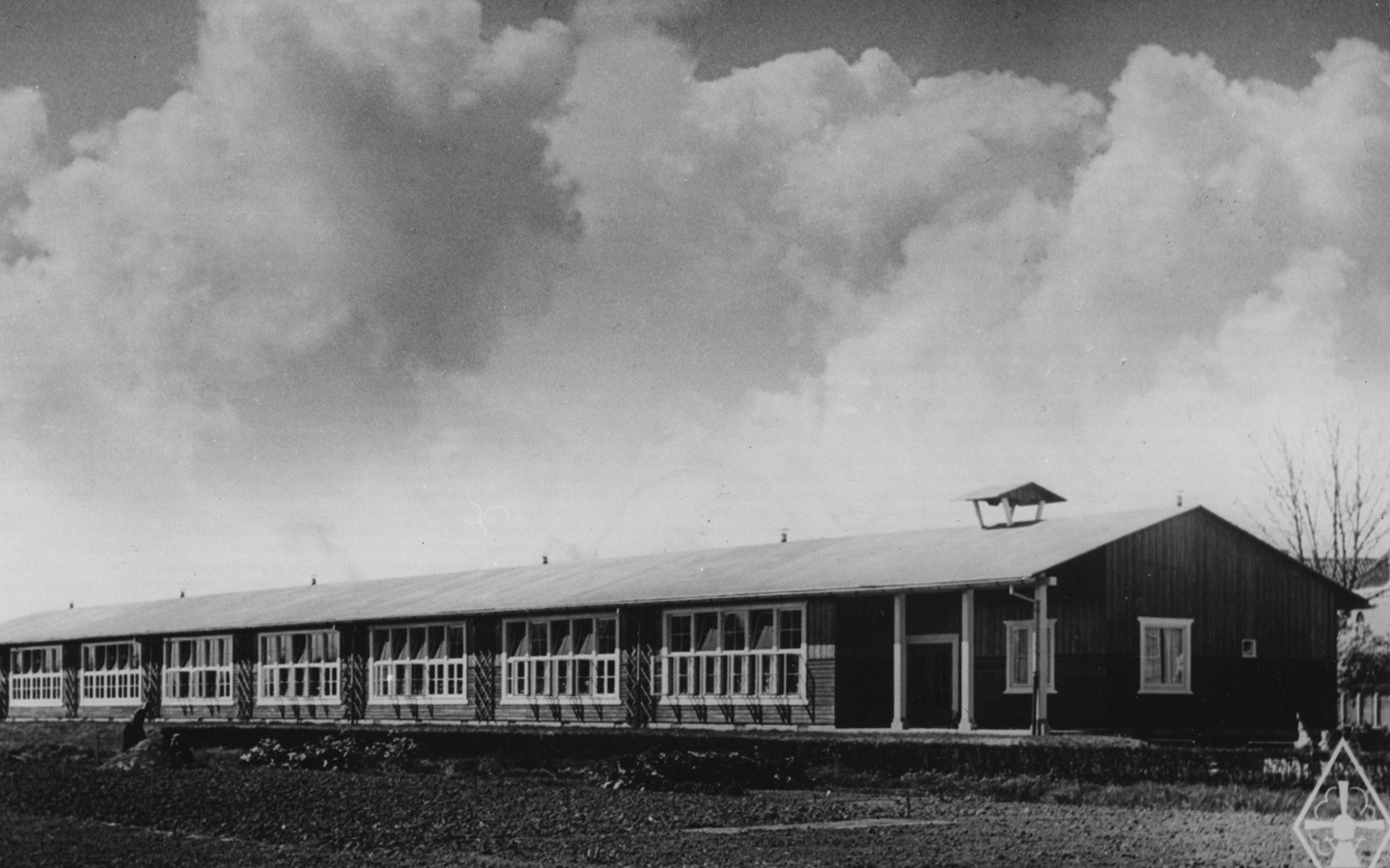 Black and white picture of a long one-storey school building with a pitched roof. Dark-painted wooden facades, white window frames. 