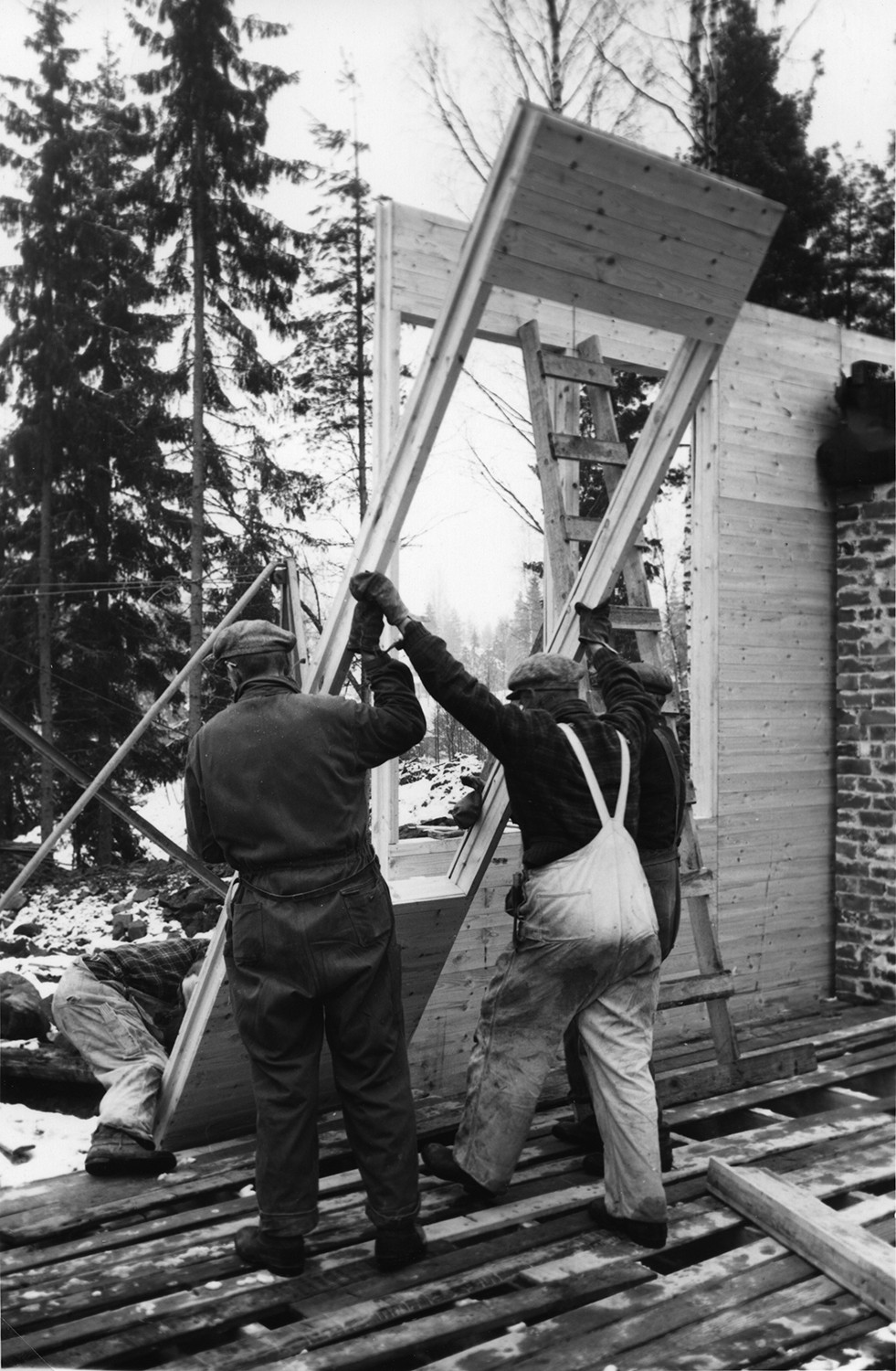 A black and white photo of three workers putting up a wall of a wooden Puutalo school.